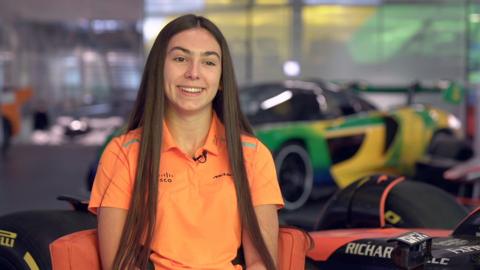 Ella Lloyd has long dark hair and is wearing a bright orange polo shirt with a McLaren logo