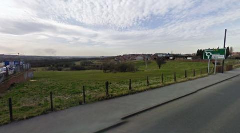 A view from an A road in Barnsley, with the road approaching a roundabout with signs towards Rotherham and Wakefield. A wire fence runs alongside the road, with a green field stretching into the distance.   