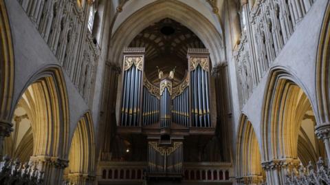 An organ is at the back of a cathedral. There is light coming in through the windows.