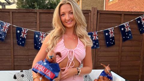 Photograph of Chanel Buckland, aged 26. She wears a pink tie crop top and a matching pink skirt. She has long blonde hair to her upper arm and smiles at the camera. She holds an inflated kangaroo that has an Australia flag around its neck. Behind her, there are Australia flags on a brown garden fence. A white garden sofa can be seen in the background. 