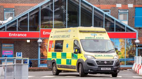 The exterior of a hospital emergency department. The building has a brick facade with a large triangular glass structure above the entrance, which is clearly marked with bright red signage reading “Entrance” and partially visible “Emergency”.
In the foreground, there is a yellow and green emergency ambulance parked near the entrance. The ambulance has the NHS logo and the words “Yorkshire Ambulance Service NHS Trust” and “Emergency Ambulance” written on it.