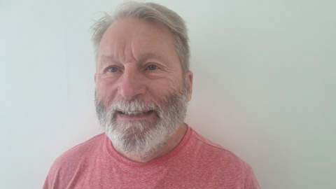 A man with grey hair and a grey beard is prominent in the photo and wearing a T-shirt. He is in front of a plain, grey background.