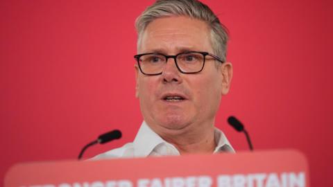 Sir Keir Starmer speaking at a podium, with Stronger Fairer Britain written along the bottom and two microphones poking out. He is wearing black rimmed glasses and an open necked shirt