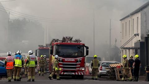 Grey smoke surrounds a fire engines and firefighters on a street
