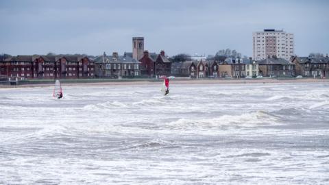 A sea coastline with two people windsurfing along the waves which are small. The weather is overcast and in the back there is houses along the seafront, and a block of flats in the corner.