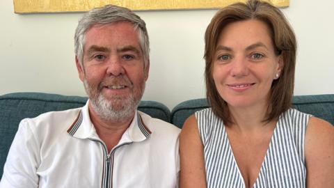 A man with short grey hair and a short grey beard with some gingery flecks wearing a white polo shirt with red and blue trim on the collar. He is sitting next to a woman with neck-length brown hair wearing a blue-and-white-striped summery dress. They are sitting on a blue sofa.