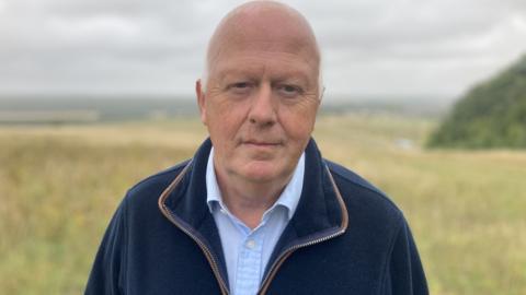 Hugh Carter standing on Salisbury Plain. he is mostly bald and wearing a blue zip-up top and light blue shirt