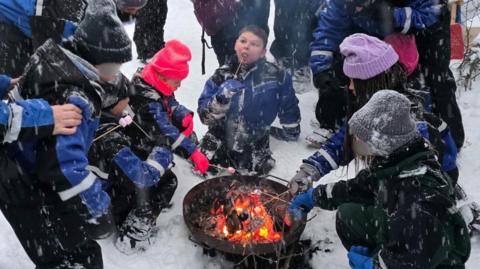 A group of adults and children sitting around a camp fire in the snow, roasting marshmallows. The children's faces are obscured so they cannot be identified.