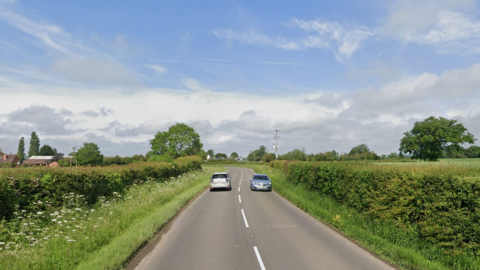 A single-lane carriageway in countryside lined with hedge rows 