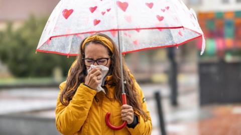 A woman blowing her nose and sneezing into a handkerchief during a rainy autumn day. She is wearing a yellow rain coat and a red umbrella