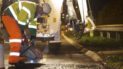 A worker in high-vis jacket and trousers, bends over a road using cutting machinery to cut into the surface. They wear a white hard hat. Behind them is a white vehicle which appears to have a hydraulic arm on it. Next to the road we can see a grass verge and a metal barrier.