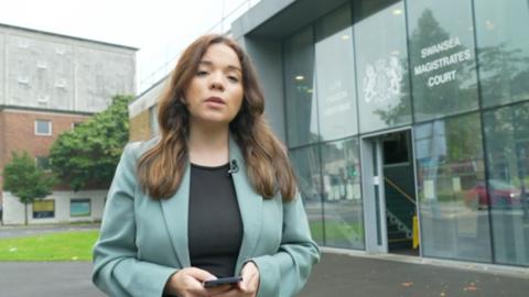 Aimee Thomas stood outside a glass windowed building which reads Swansea Magistrates Court. She's got long brown hair and is wearing a teal blazer and black top. She is holding a mobile phone