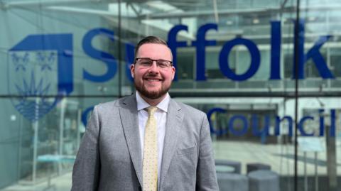 Michael Hadwen is standing in front of a glass building at Suffolk county council. He is wearing a grey suit, white shirt and yellow tie.