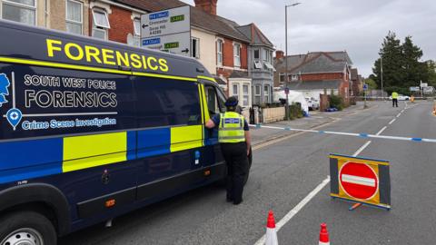 A navy blue police forensics van waits at a police tape cordon on a residential street. The van has forensics written in yellow reflective writing and South West Police Forensics written in blue writing on a black tinted window. An officer in a hi-vis vest stands at the window of the van