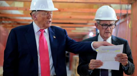 US President Donald Trump and Federal Reserve Chair Jerome Powell tour the Federal Reserve’s headquarters renovation project on July 24, 2025. Both men are wearing white hard hats, and suits, as Trump points at a piece of paper Powell is holding, which both men are looking at.