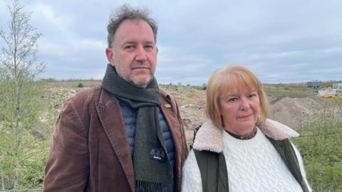 Chris Green and Liz Reid standing near piles of rubble at the quarry site. Chris has short grey hair, a brown tweed jacket and scarf and a blue jumper. Liz has short blonde hair with fringe and wears a white jumper and a black body warmer.