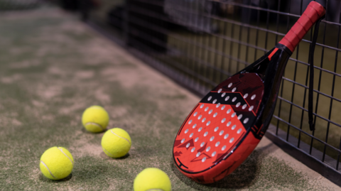 A red and black padel racquet near four yellow balls on a green court 