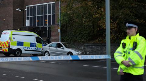 A police officer stands in front of police tap which covers the synagogue. There is a police van behind the cordon and a silver car.
