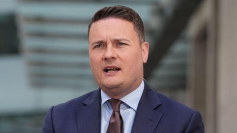 Wes Streeting is dressed in a blue suit and brown tie. He has combed, brown hair which is shaved at the sides.