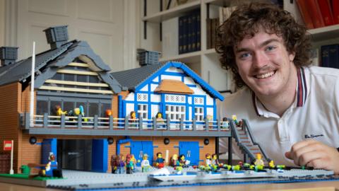 Jim Gleed is sitting by a Lego model of a boathouse. He has curly brown hair and a moustache, and is wearing a white polo shirt. The Lego model shows the ground floor and first floors of the boathouse which is mainly blue and white. Lego figures can be seen on the decks and in front of a river.