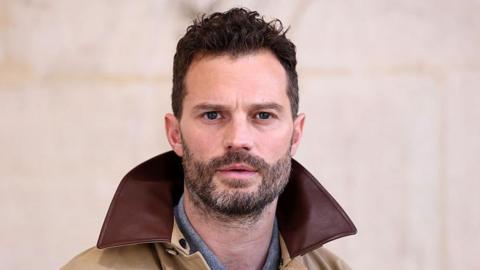 Jamie Dornan is wearing a beige jacket with a brown collar, and looks directly at the camera.