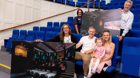 In a lecture theatre with royal blue chairs, three adults are sat in the front row, a child is on the lap of the woman on the right of the picture. The woman on the left of the row is holding a big board showing a CGI image of a cinema. Behind them a man and a woman are holding a board together, it shows the cinema from a different angle