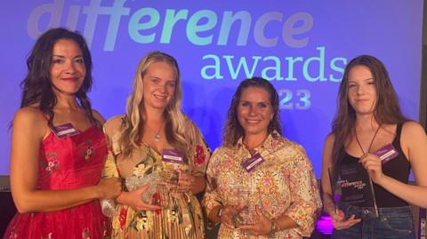 Four women at an award ceremony are wearing smart frocks and smiling for the camera 