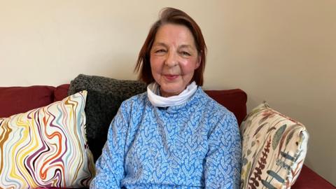 Hazel has brown hair and looks to camera whilst sitting on her sofa in a blue top, She has a white neck tie on.