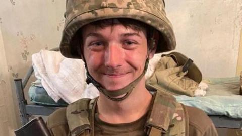 James Witton smiles for the camera, he is wearing a military helmet with camouflage covering. Behind him is a bed with a bare mattress and some objects on it   