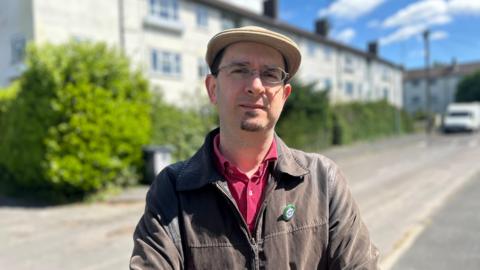 Head and shoulders image of Maurice Chiodo on Ekin Road in Cambridge. He is wearing a flat cap, thinly rounded glasses and has a goatee. He is wearing a brown jacket and red shirt.