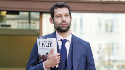 Adam Aspinall Da Encarnacao carrying a copy of the book Credible And True: The Political And Personal Memoir of K Harvey Proctor