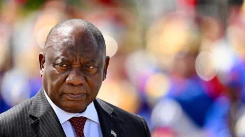 A shot of President Cyril Ramaphosa, wearing a pin striped black suit with a white shirt and red patterned tie, from the shoulders up. He is looking off to the left and the background is blurred.