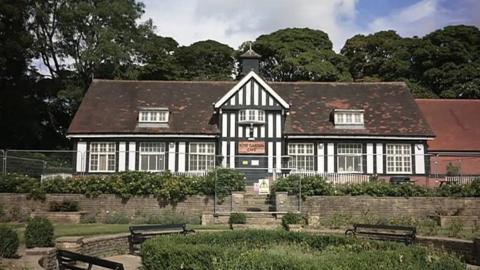 A building with black and white Tudor style panels has wooden windows and a pitched roof. It overlooks a garden with benches