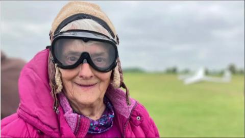 Barbara with flying goggles on smiling to camera with airfield in background.