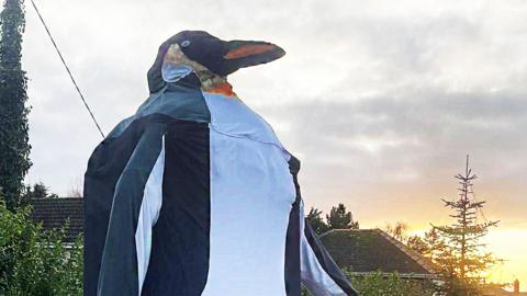 The head and shoulders of the large penguin artwork. It is made from black and white fabric and has an orange and black beak. It is pictured against a cloudy sky.