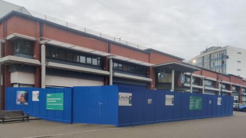 The entrance to Barrow Market Hall. The long two-storey redbrick building has a sign above the doorway and is surrounded by blue wooden boards. Street lights can be seen on the flat roof.