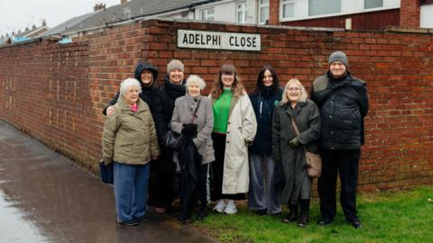 A group of seven women and one man stand in the rain on a residential street. The street sign above their heads reads Adelphi Close.