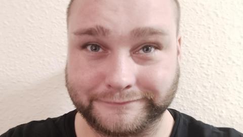 A close-up selfie of a man with a light brown beard and light-brown eyes. He is smiling at the camera. There is a white wall behind him.