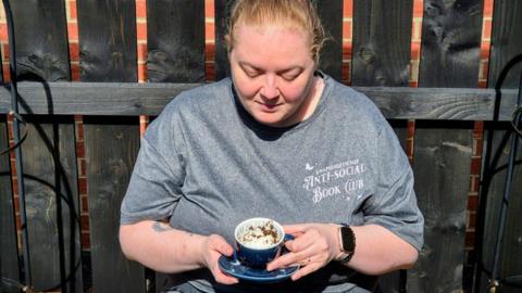 Michelle Liddle is sat in front of a wooden fence tilting a blue teacup to show tea leaves inside. She is wearing a grey t-shirt and her blonde hair is tied back.