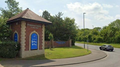 A small red brick tower with roof stands on grass, beside a pavement. It says Primrose Valley Holiday Park and there is a blue logo beneath the sign. To the right, a low brick wall leads away from the tower.