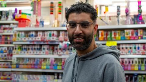 A man with a beard and wearing glasses stands in front of rows of colourful vapes on the shelves in a shop