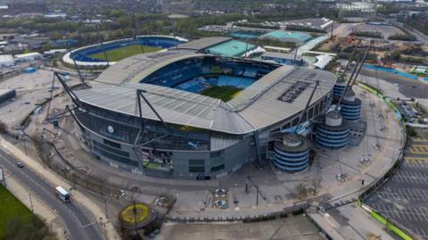 A large football stadium see from above. It is surrounded by a carpark on one side, another football pitch on another and a road on another. It has the word Etihad printed on the top of the main stadium. 