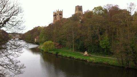 Shot of a curving river with trees and green grass on the far side. In the background you can see a large cathedral - Durham Cathedral. The water is clear and reflecting the grey sky.