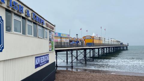 A pier stretches out to sea with flags and attractions on its flanks and metal structural legs.