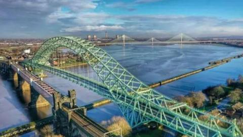 A green metal bridge loops in the centre and comes down across the River Mersey