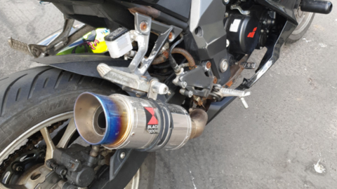 The picture shows the rear right-hand side of a black sports motorcycle parked on a road surface. The most noticeable feature is an aftermarket exhaust canister with a blue-tinted metal tip and a visible "Black Widow" logo, along with some rust and wear around parts of the frame and exhaust system.