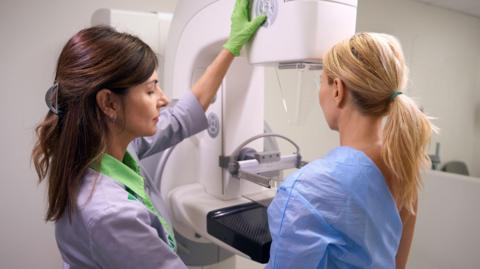 A woman stands in a hospital gown undergoing a mammogram. A medical practitioner stands beside her in a black top.