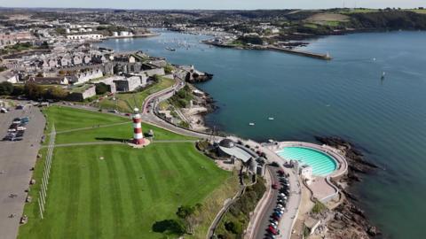 A view of Plymouth Hoe and the city seafront. It features red‑and‑white striped Smeaton’s Tower lighthouse, and a large green lawn around it, which is Plymouth Hoe Park. The Tinside Lido, a curved open‑air seawater pool built into the rocks is on the right. A harbour and breakwater can be seen in the distance.