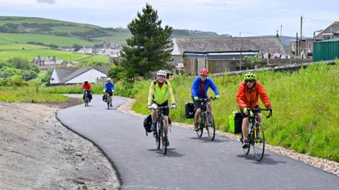 Five cyclists using the revamped cycle path in Lowca. A group of three is in the foreground, with two cyclists a short distance behind them. There are houses on the right hand side and in the distance, surrounded by lush green countryside. All the cyclists are wearing helmets and colourful jackets, some have bags attached to their bikes.