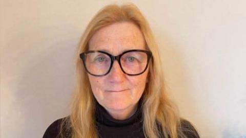 An elderly woman, blonde, with thick black glasses standing against a wall wearing a black polo neck top